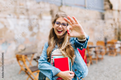 Charming female student with books showing her excitement and holding up her thumb. Outdoor portrait of young lovely lady with smiling facial expression.