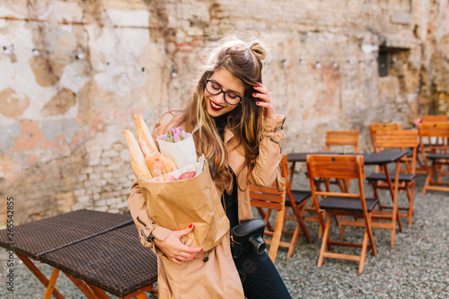 Nice shy girl with long hair looks at the bakery bag standing in outdoor restaraunt in front of old building. Pretty stylish lady in glasses straightening her hair and posing after food shopping.