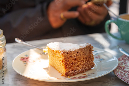 Close up view on a piece of cake with icing sugar, on the white plate beside sugar bottle and cup of coffee, and outdoor vintage sunshine atmosphere with blur background people use mobile phone.