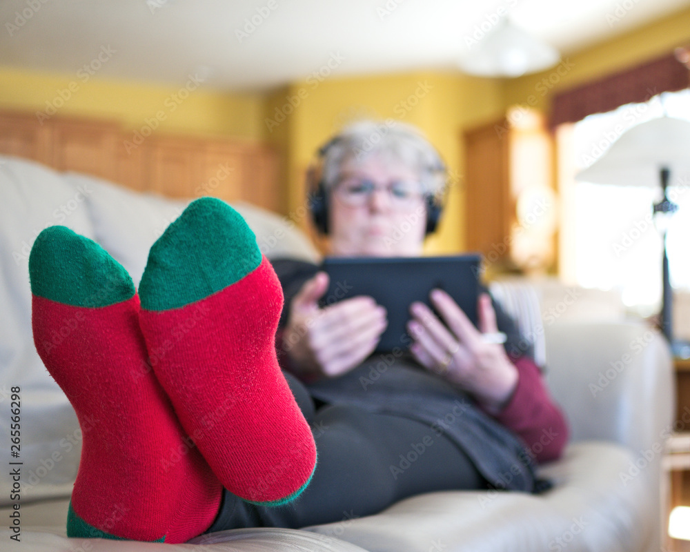 Obraz premium Senior woman with red socks relaxing on sofa at home with tablet and headphones. Grandmother reading and listening to music with her feet up and toward camera.