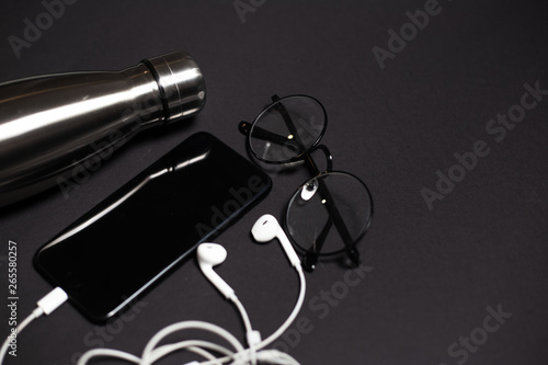 Top view of black desk with steel thermo bottle, glasses, smartphone and earphones.