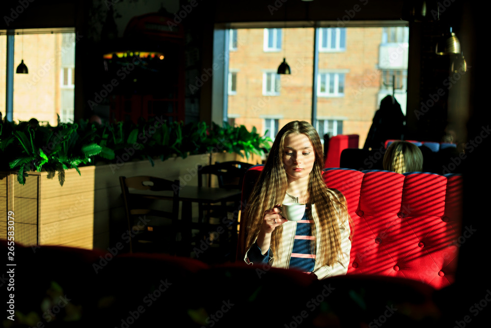 Young pretty white girl in casual clothing sits on a red couch and drinks coffee near a window in a cafe. Shade from the blinds. Thin lines of light on the hair.