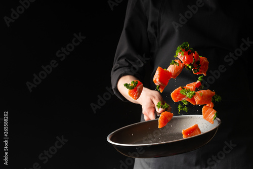 Fototapeta Naklejka Na Ścianę i Meble -  Sea cuisine, Professional cook prepares pieces of red fish, salmon, trout with greens. Frost in the air, Cooking seafood, healthy vegetarian food and food on a dark background. Horizontal view. Banner