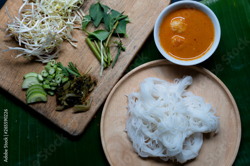 Rice noodles in fish curry sauce with vegetables on a wooden plate and place on the banana leaf floor. Separate coconut milk curry with meat ball and vegetables.