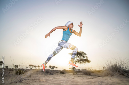 Side view of a strong African Black woman with long braids in hair in the desert in sportswear does a dramatic running leap against a golden sunset or sunrise holding a bamboo stick 