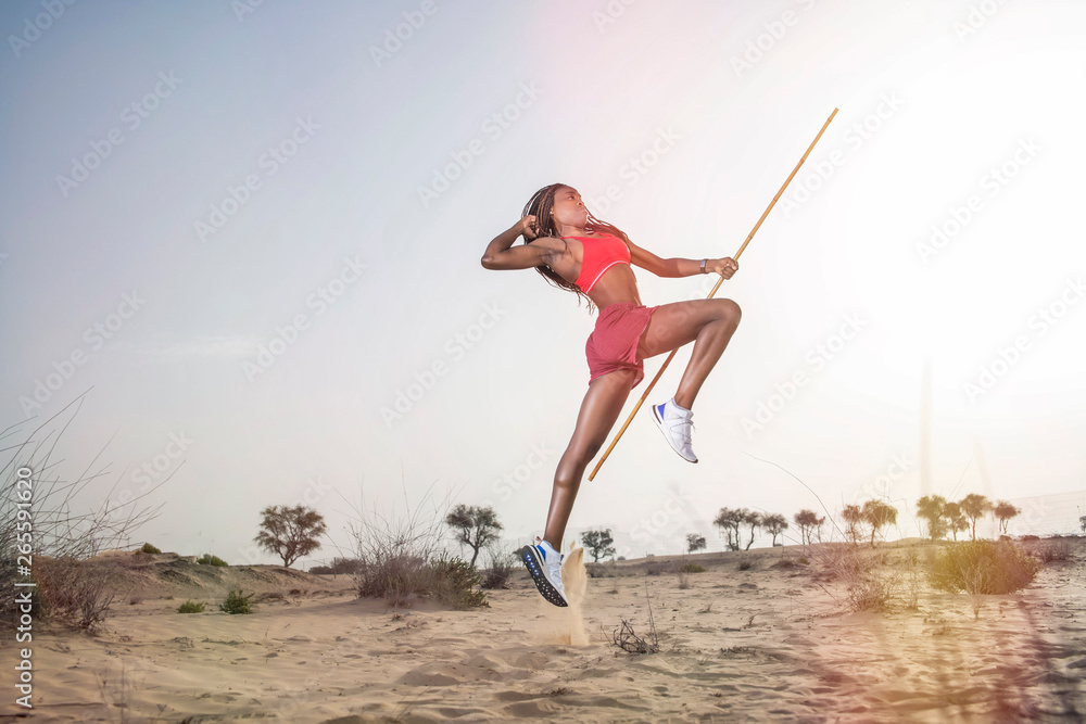 Side view of a strong African Black woman with long braids in hair in the middle eastern desert in sportswear does a dramatic running leap against a golden sunset or sunrise  holding a bamboo stick   