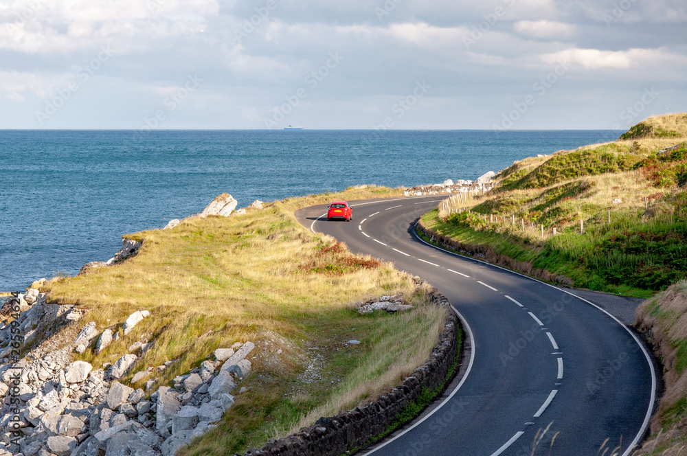 The eastern coast of Northern Ireland and Antrim Coastal Road, a.k.a. Causeway Coastal Route with a red car. Sunset light