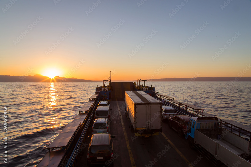 Ferry car and truck transportation in an idyllic sunset back lighted tranquil scene with orange reflection in water and sunbeams 