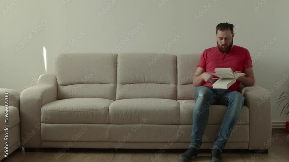 Caucasian beard man sitts on a sofa inside a room and reading a book. He doesnt know where to start the reading again and smilles Caucasian beard man sitts on a sofa inside a room and reading a book. He doesnt know where to start the reading again and smilles