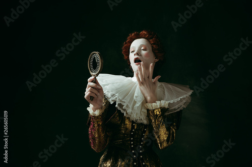 Self loving. Medieval redhead young woman in golden vintage clothing as a duchess looking in the mirror on dark green background. Concept of comparison of eras, modernity and renaissance.