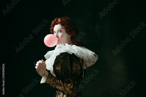 Medieval redhead young woman in golden vintage clothing as a duchess with red sunglasses blowing a bubblegum on dark green background. Concept of comparison of eras, modernity and renaissance. photo