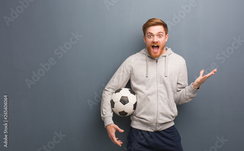 Young redhead fitness man celebrating a victory or success. He is holding a soccer ball.