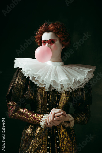 Fotografie Medieval redhead young woman in golden vintage clothing as a duchess with red sunglasses blowing a bubblegum on dark green background