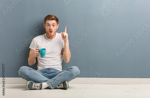 Young redhead student man sitting on the floor having a great idea, concept of creativity. He is holding a coffee mug.