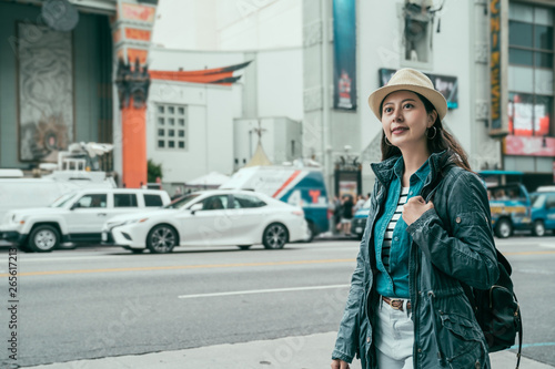 Asian woman travel backpacker vivacious tourism in los angeles standing on hollywood walk of fame with beautiful beaming smile on busy street. Chinese theatre old monument in background in urban.