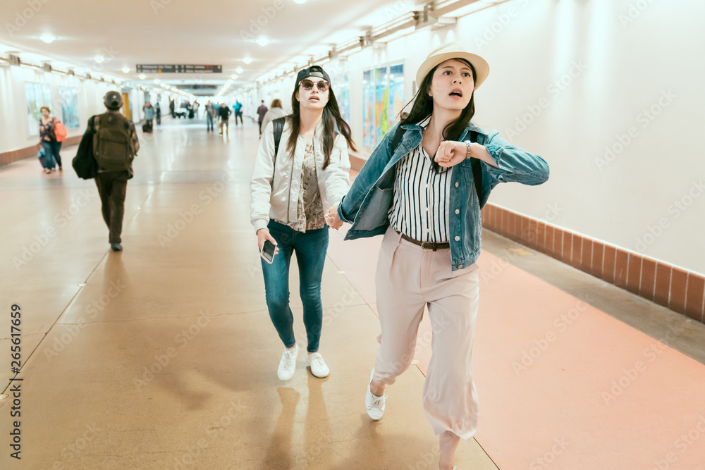 Full length portrait of nervous outgoing asian girls friends with phone in hand hurrying at airport underpass. two women travelers running late for train watching time rush in corridor union station