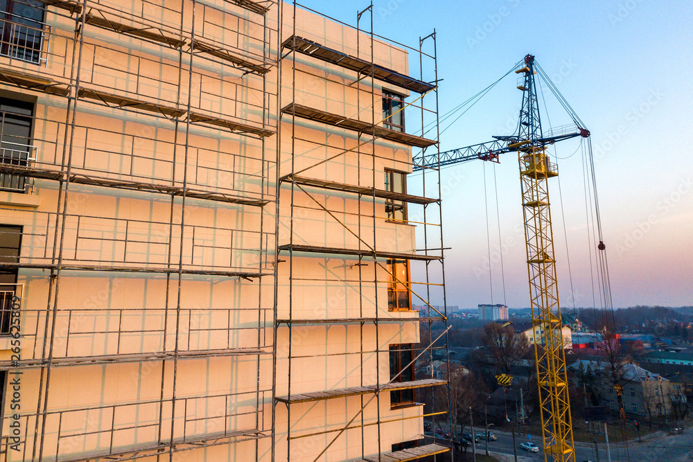 Apartment or office tall unfinished building under construction. Brick wall in scaffolding, shiny windows and tower crane on urban landscape and blue sky background. Drone aerial photography.