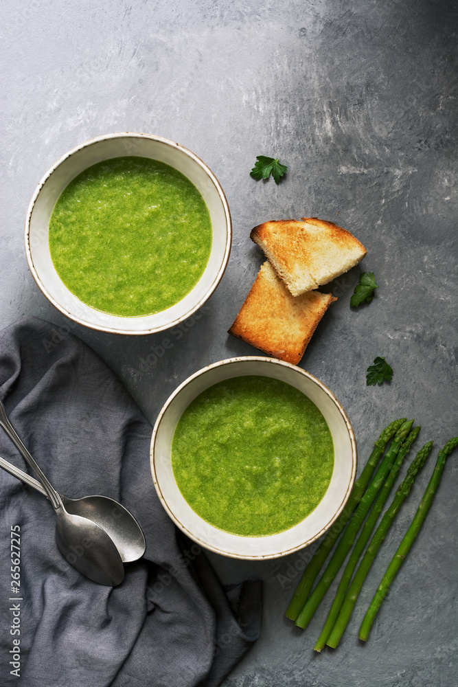 Two bowls of green cream asparagus soup with crispy toasts on a gray background. Overhead view, copy space. Two bowls of green cream asparagus soup with crispy toasts on a gray background. Overhead view, copy space.