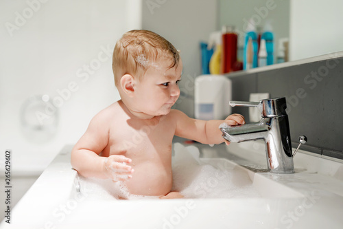 Cute adorable baby taking bath in washing sink and playing with water and foam