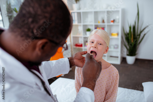 Dark-skinned doctor examining throat of schoolgirl feeling bad