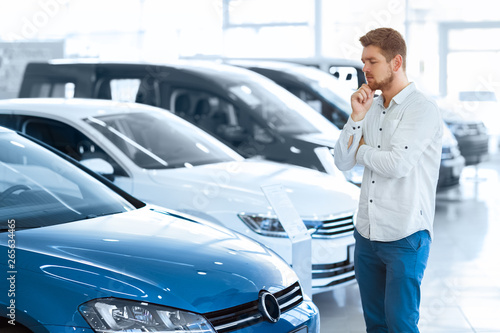 Final decision. Shot of a handsome young man standing in front of a new car at the dealership thinking rubbing his chin 