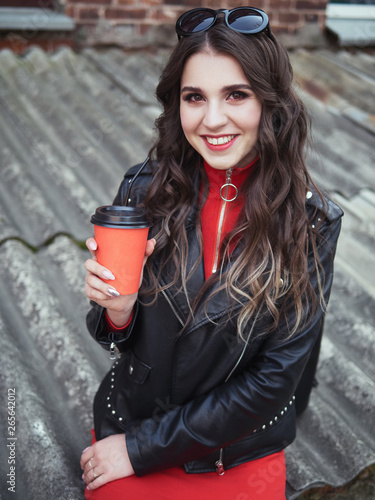 Young Adorable Brown Haired Teen Lady With Bright Trendy Makeup Adorable Smile In Red Dress Black Leather Jacket Holding Red Coffee Cup Outdoors Enjoying Her Vacations Sitting In Old Town Courtyard Stock