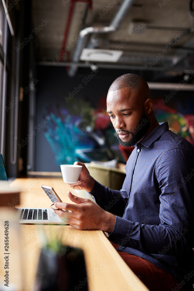 Fototapeta premium Mid adult black male creative sitting by window having coffee using smartphone, side view, vertical