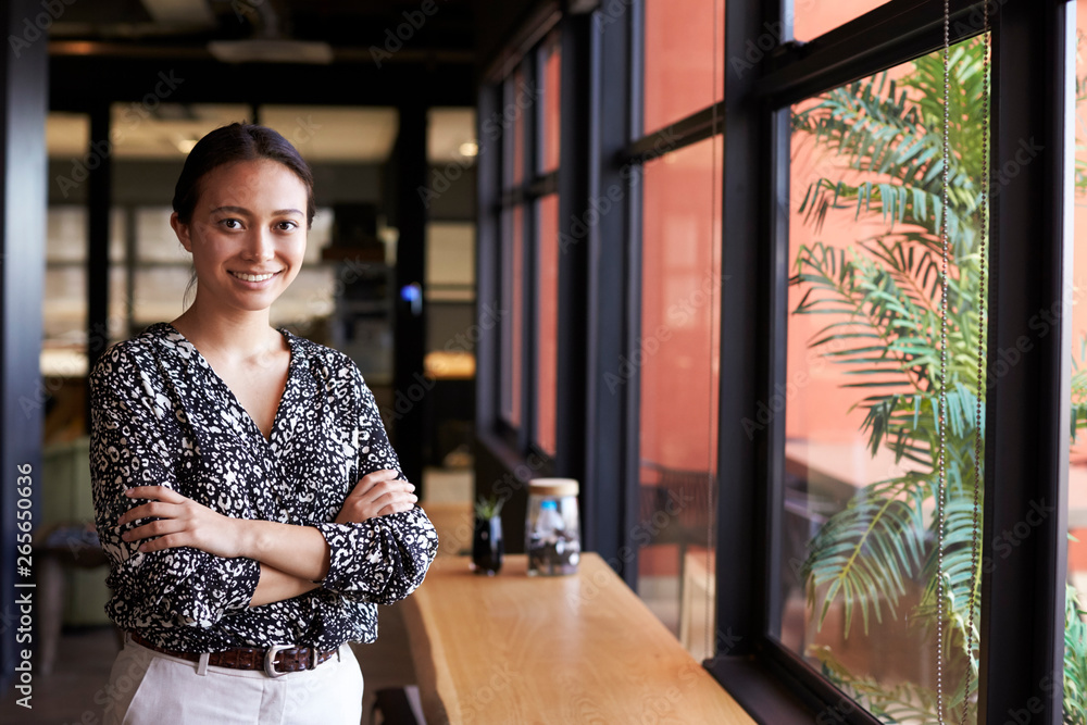 Millennial Asian businesswoman standing by the window in an office smiling to camera, waist up