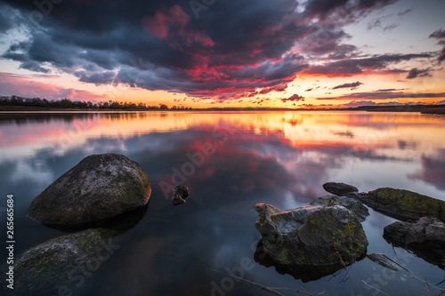 Feuriger Sonnenuntergang über dem Sehlendorfer Binnensee mit Steinen im Vordergrund und Reflektionen von Wolken im Wasser, Schleswig-Holstein photo