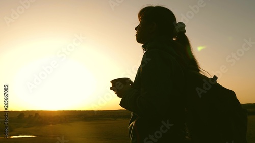 silhouette woman traveler, stands on top of hill drinking coffee in glass from thermos. tourist girl drinks hot tea and looks at sun. Adventurer rest after reaching goal. freedom concept.