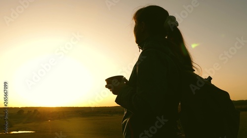 silhouette woman traveler, stands on top of hill drinking coffee in glass from thermos. tourist girl drinks hot tea and looks at sun. Adventurer rest after reaching goal. freedom concept.