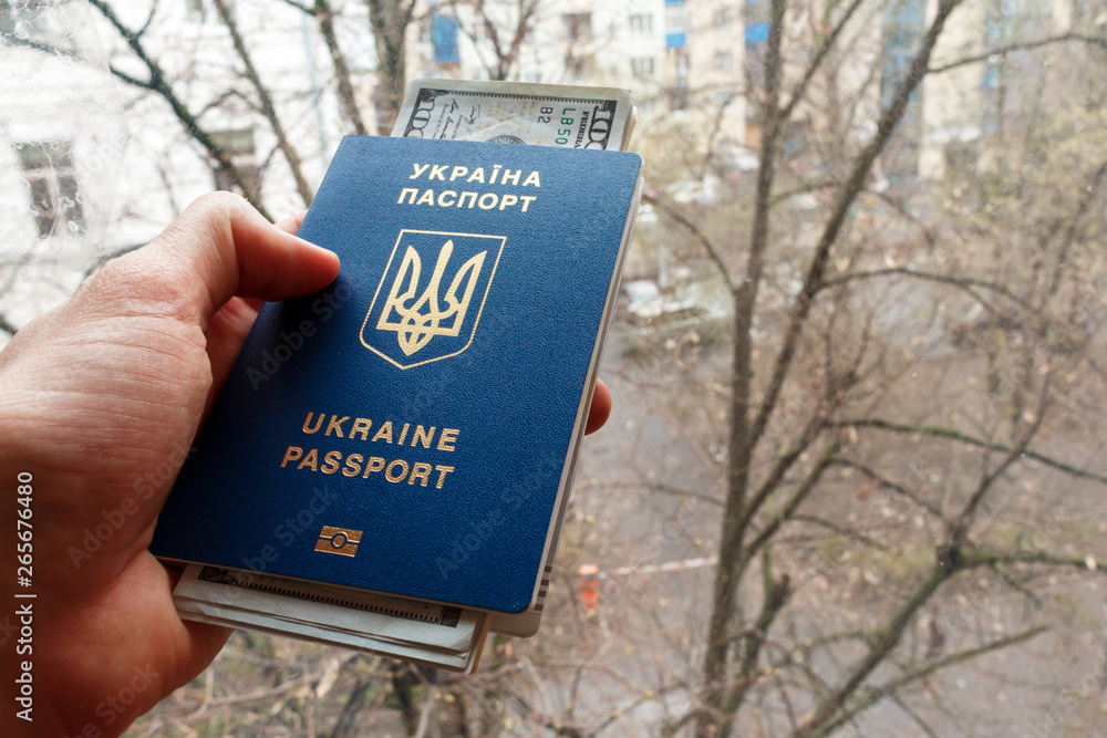 Ukraine passport in hand with USA dollars against the background of the window, close-up. Migration from Ukraine and Travel concept. Ukrainian blue International biometric passport