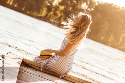 Back view of beautiful blonde girl in stripped overalls sitting on wooden pier with her hair in motion. Attractive young woman in stylish summer clothing posing with fluttering hair at sunny riverside