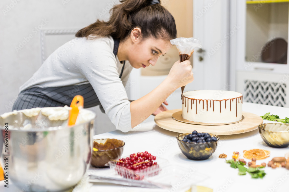 A confectioner squeezes liquid chocolate from a pastry bag onto a white cream biscuit cake on a wooden stand. The concept of homemade pastry, cooking cakes. A confectioner squeezes liquid chocolate from a pastry bag onto a white cream biscuit cake on a wooden stand. The concept of homemade pastry, cooking cakes.