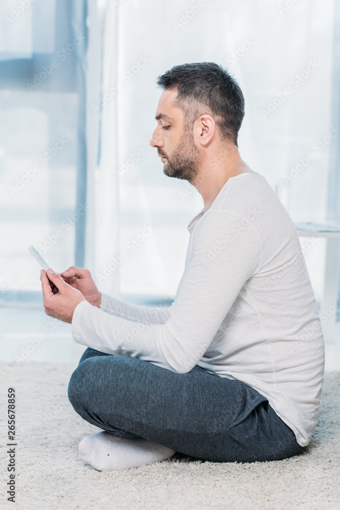side view of handsome man in casual clothes sitting on carpet and using smartphone at home side view of handsome man in casual clothes sitting on carpet and using smartphone at home