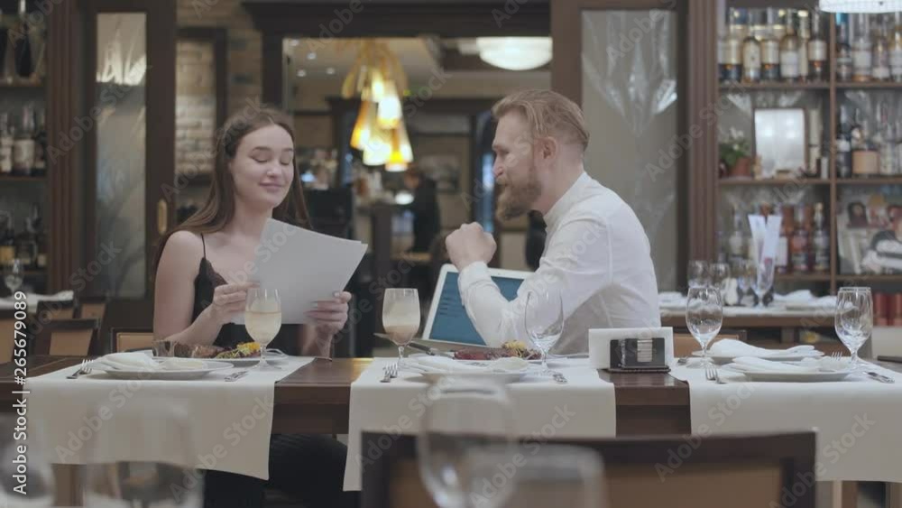 Portrait cute young woman sit with papers on the table in restaurant with a confident bearded man who show her information on the laptop. Business dinner. Portrait cute young woman sit with papers on the table in restaurant with a confident bearded man who show her information on the laptop. Business dinner.