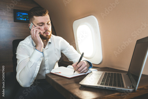 Young attractive and successful businessman talking on the phone and working while sitting in the chair of his private business plane