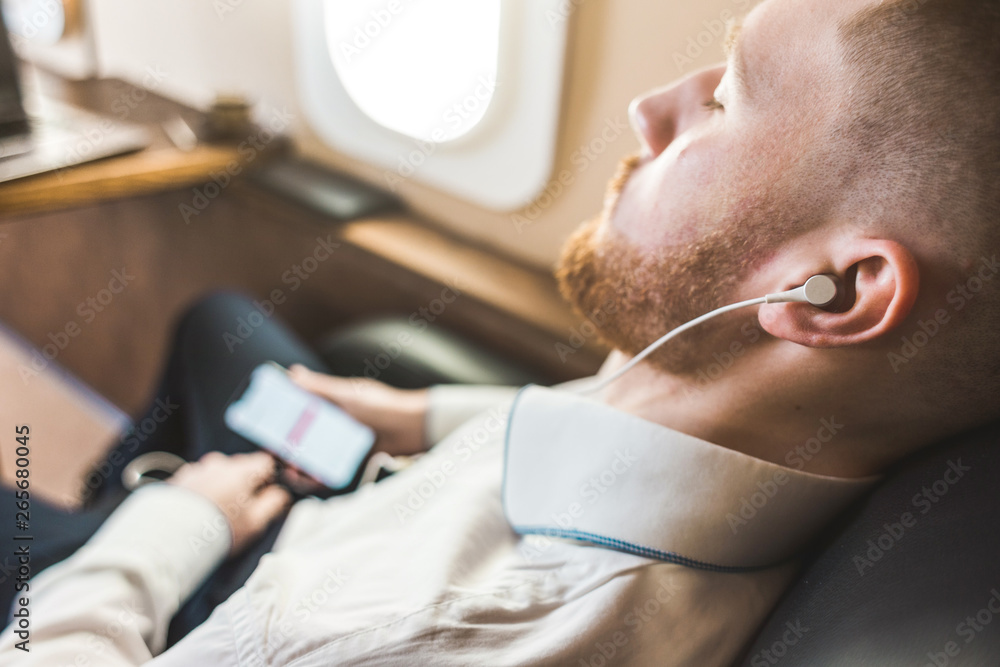 Fototapeta premium Young attractive and successful businessman is resting and listening to music works while sitting in the chair of his private business plane
