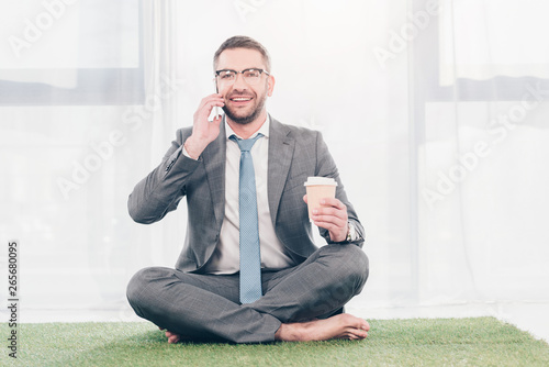 handsome smiling businessman in glasses and suit sitting on grass mat with coffee to go and talking on smartphone