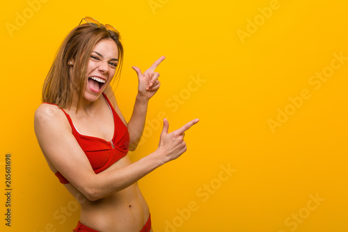 Young caucasian woman wearing bikini and sunglasses pointing with forefingers to a copy space  expressing excitement and desire.