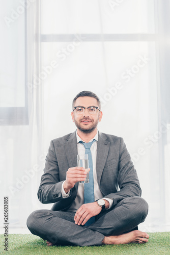 handsome businessman in suit sitting on grass mat, looking at camera and holding glass of water