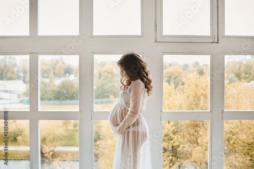 Beautiful young pregnant woman with fashionable hairstyle in a stylish peignoir smiling and posing near the big window at light interior