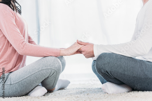 cropped view of man and woman in casual clothes sitting in lotus pose, meditating and holding hands at home