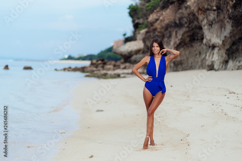 Slender sexy tanned woman in blue swimsuit posing on beach with white sand