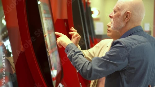 Senior couple choosing movie and buying ticket from vending machine at movie theater at mall via touch screen. Senior man making gestures by touching screen. Active modern life after retirement.