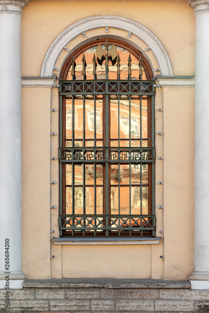 A window with an arch and a lattice with peaks on a yellow wall between two white columns. Old European architecture