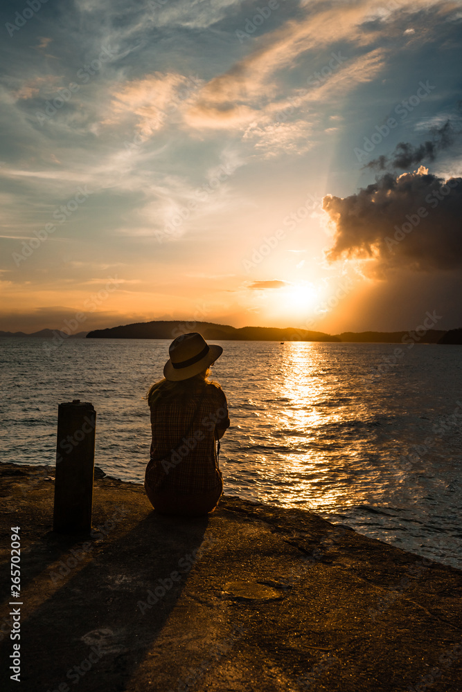 Fototapeta premium silhouette of a Girl in straw hat sitting in front of ocean and sunset cloudy blue and orange sky