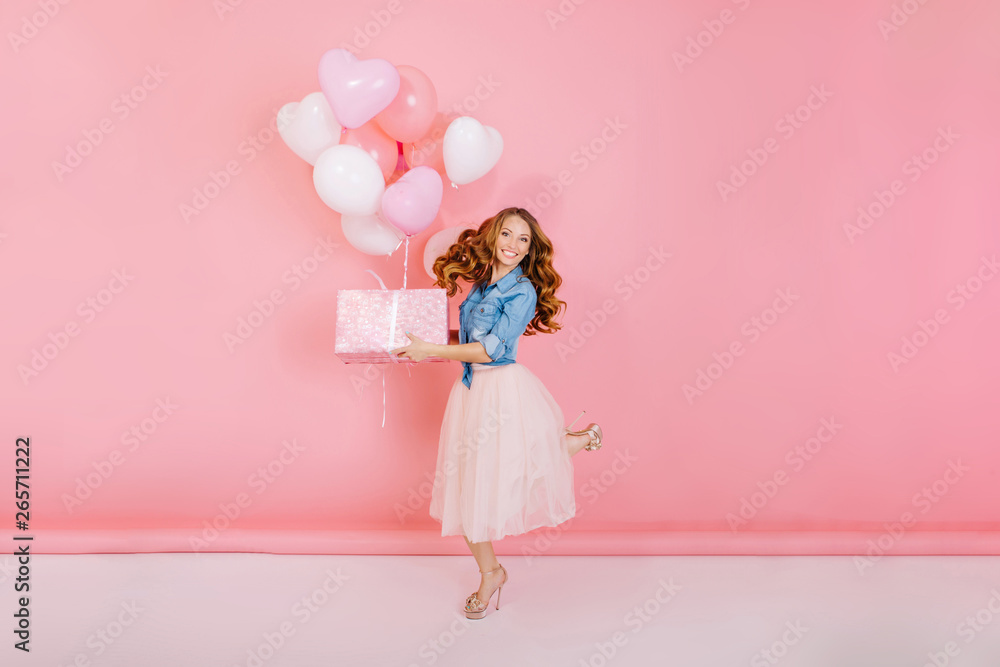 Full-length portrait of adorable laughing birthday girl, jumping with present box and balloons. Gorgeous young woman with curly hair having fun on party holding gift, isolated on pink background.