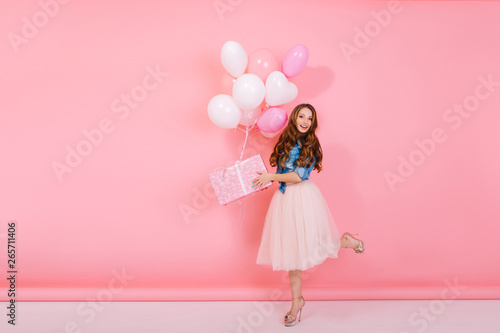 Full-length portrait of adorable birthday girl with curly hair wearing high heel shoes, holding present in cute box. Graceful slim young woman received gift with colorful helium balloons on holiday