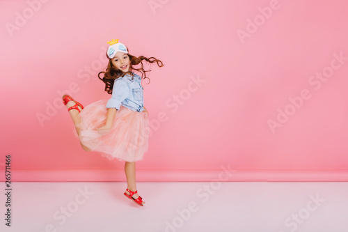 Excited joyful amazing girl with long brunette hair, in princess mask on head jumping in tulle skirt isolated on pink background. Having fun of happy kid, expressing positivity. Place for text